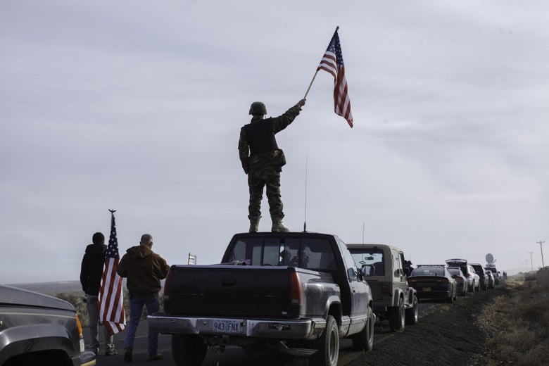 A protester dressed in military gear stands on top of a pickup truck holding an American flag.